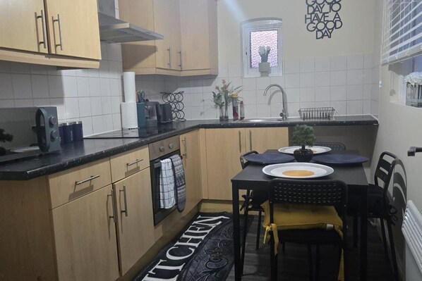 A compact kitchen with wooden cabinets, a black countertop, and a dining area with a table and chairs. - 2 Bed - Sleeps 6 - Private Balcony - City Centre (Wolverhampton)