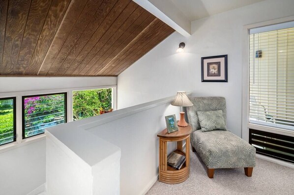 A modern room with a sloped ceiling, a window with blinds, a small wooden side table with a lamp, and a patterned chair.