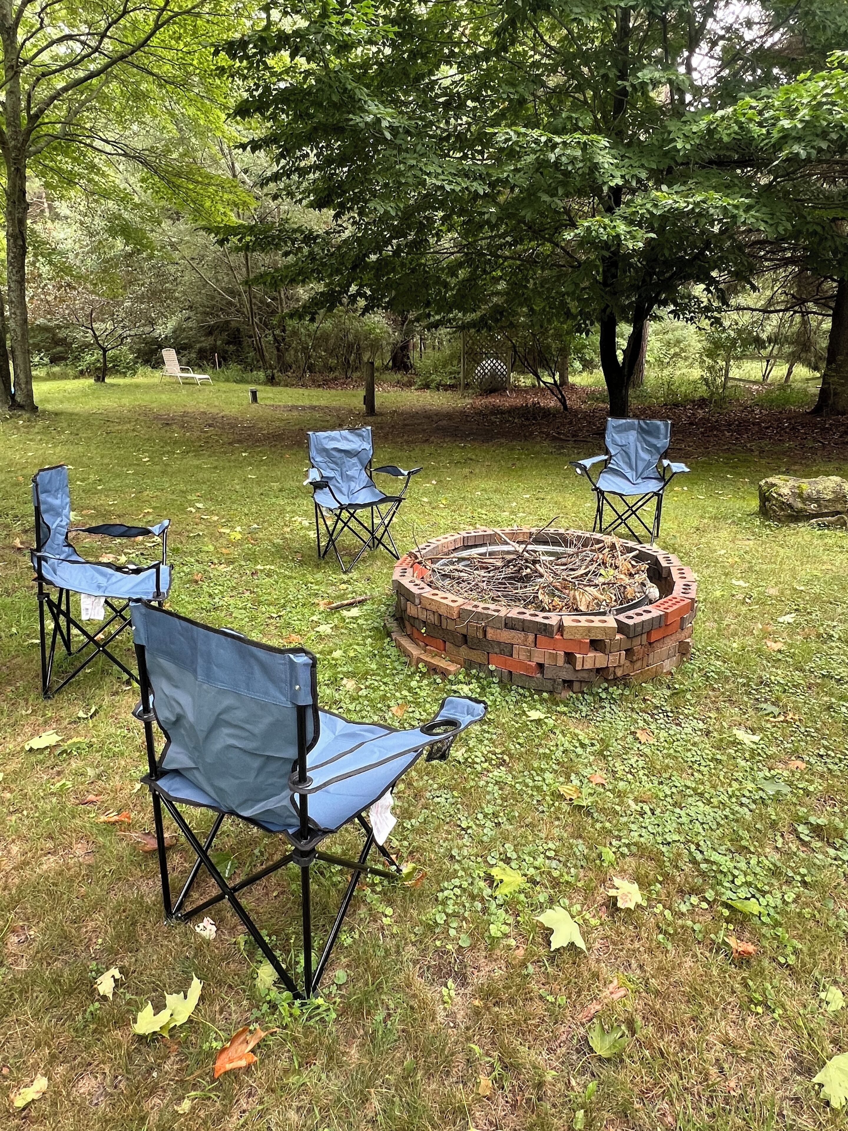 A circular brick fire pit surrounded by four blue folding chairs in a grassy outdoor area with trees.