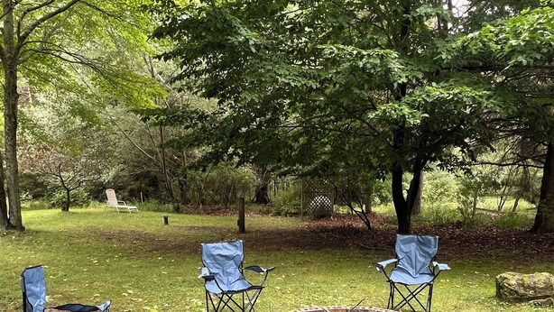 Un foyer circulaire en briques, entouré de quatre chaises pliantes bleues, se trouve dans un espace extérieur verdoyant, agrémenté d’arbres.