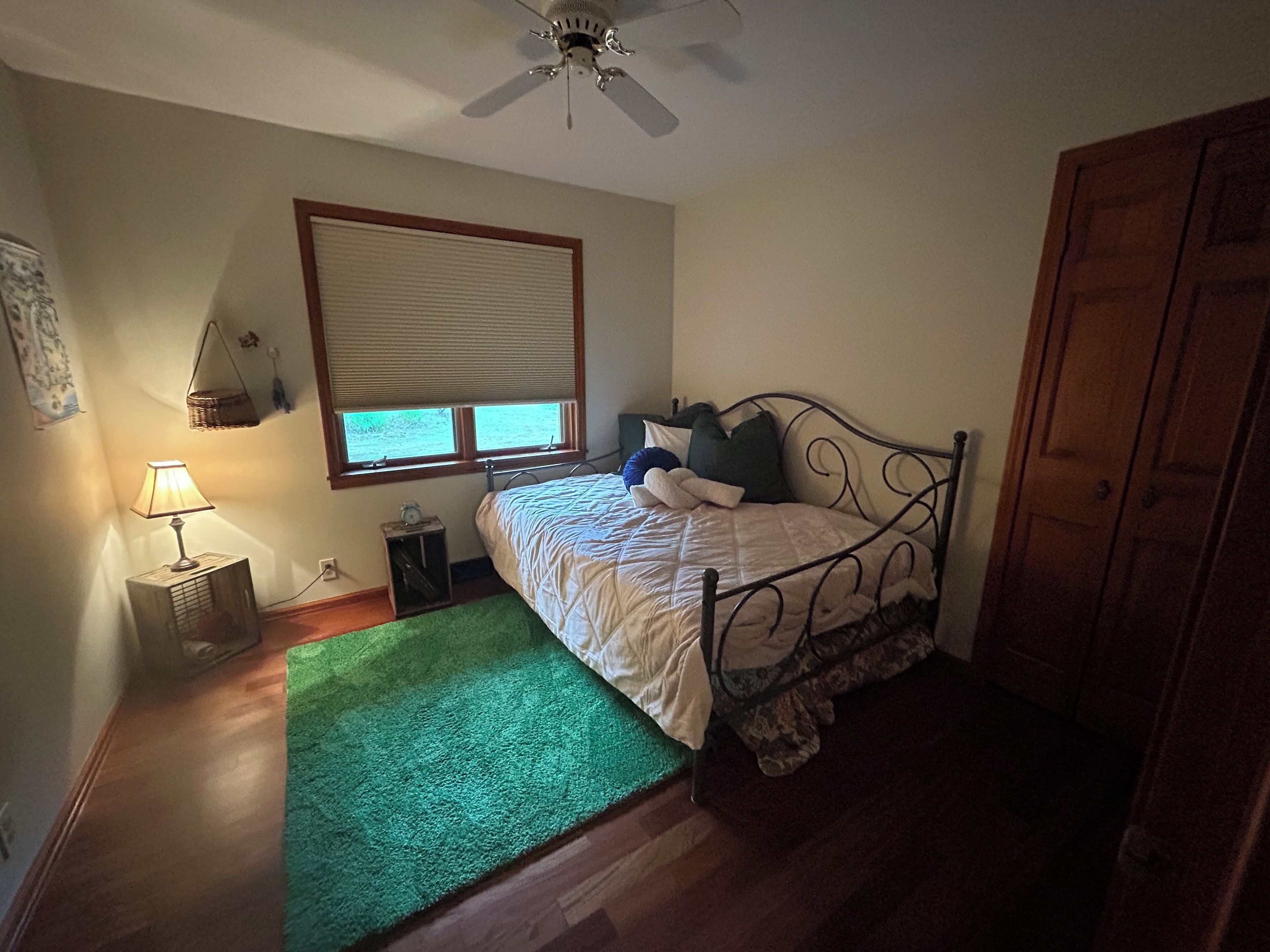 Een slaapkamer met een bed, een groen vloerkleed, een nachtkastje met een lamp, een raam met jaloezieën en een plafondventilator.