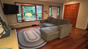 A living room with a grey sofa, wooden flooring, a ceiling fan, a TV mounted on the wall, and a window with blinds.