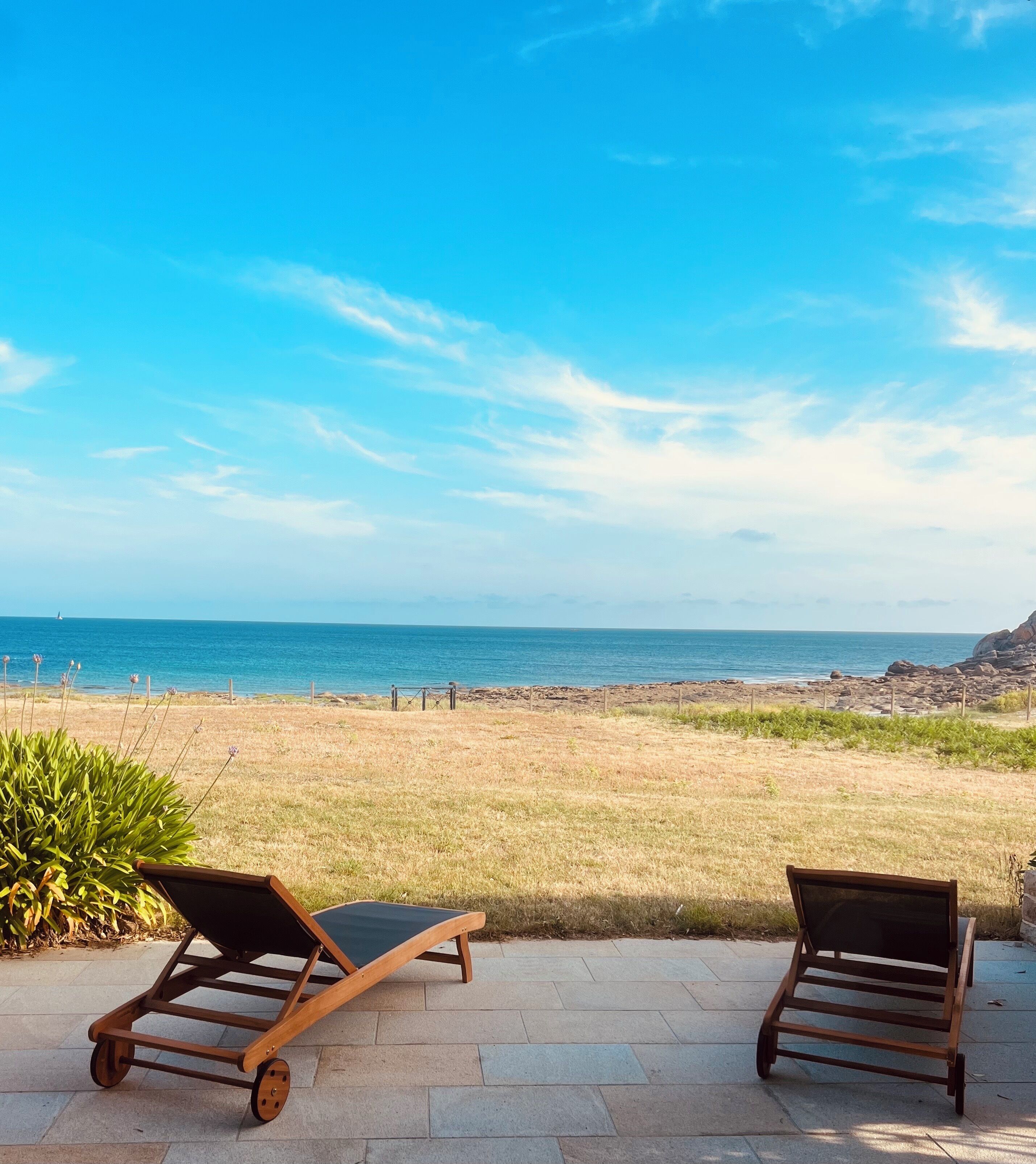 Deux fauteuils de jardin en bois, placés face à une plage, offrent une vue dégagée sur l’océan et une côte rocheuse.