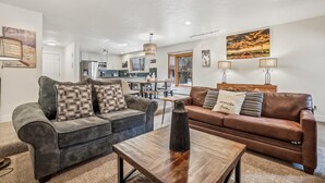 Una sala de estar con un sofá seccional gris, un sofá de cuero marrón y una mesa de centro de madera. Se observa una cocina al fondo, con un ventanal grande.