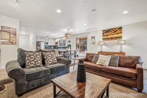A living room with a gray sectional sofa, a brown leather sofa, and a wooden coffee table. There is a kitchen in the background with a large window.