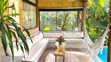 Terraza de madera con una hamaca, sillas de mimbre y un área de estar acolchada con vistas al exterior.