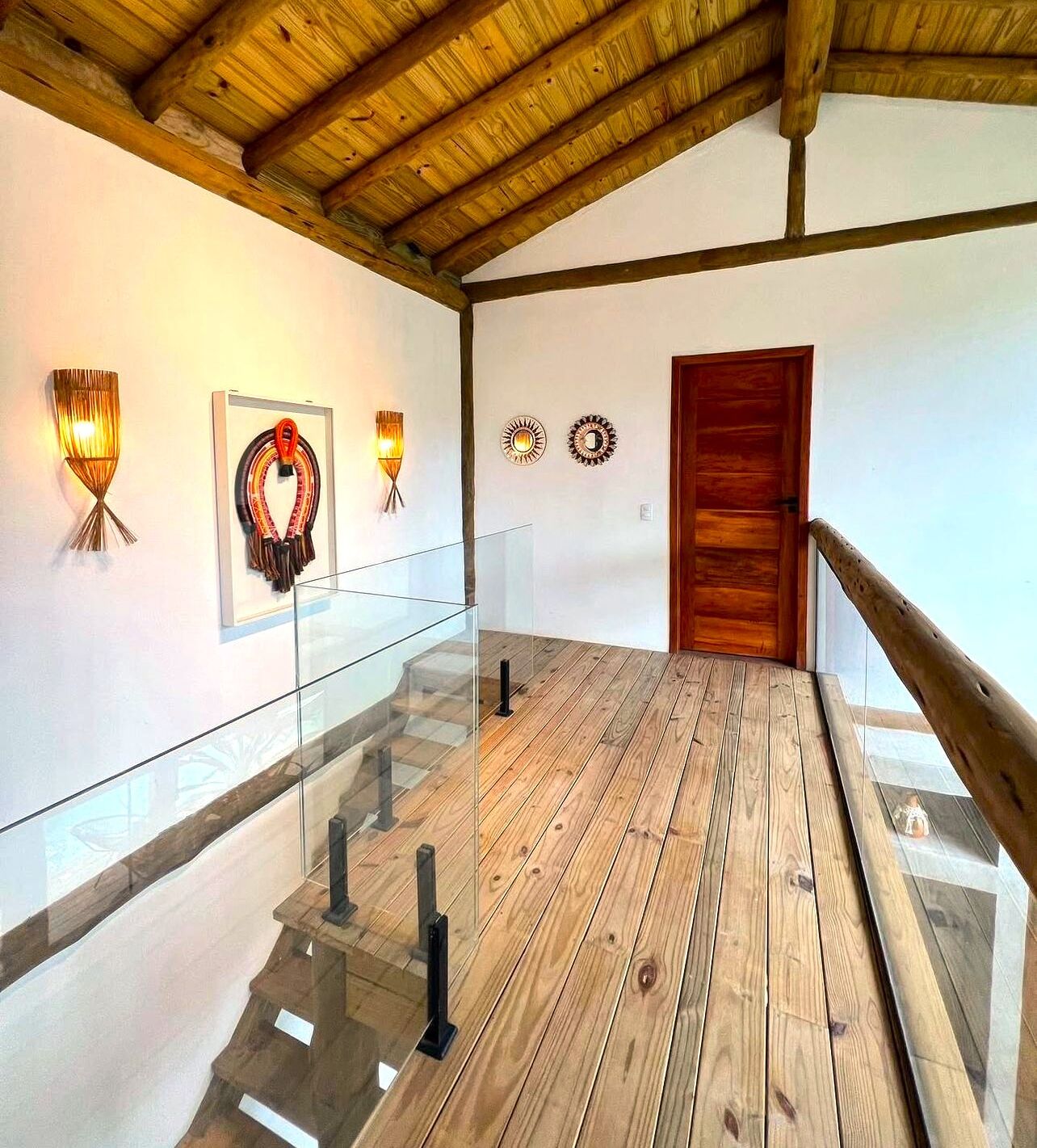 Un pasillo con suelo de madera, barandillas de vidrio y apliques de luz en la pared.