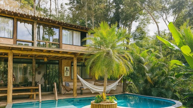 Une maison en bois à deux étages, dotée d’une piscine, entourée d’une végétation luxuriante et agrémentée d’un hamac.