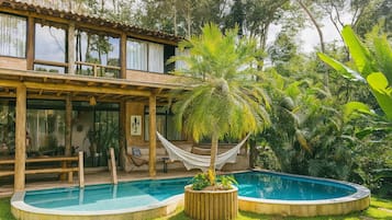 Una casa de madera de dos pisos con piscina, rodeada de exuberante vegetación y una hamaca.