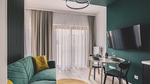 Ein modernes Wohnzimmer mit einem grünen Sofa, einem gelben Überwurf und einem runden Teppich. Es gibt einen Essbereich mit Tisch und Stühlen, und ein an der Wand befestigter Fernseher.
