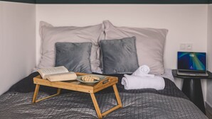 Ein ordentlich bezogenes Bett mit einem Tablett, das ein Buch und Kekse hält, Kissen, ein Laptop auf einem Nachttisch und ein Fenster mit Vorhängen.