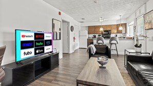 A modern living room with a flat-screen TV displaying streaming services, a wooden coffee table, a leather sofa, and a kitchen in the background. - Spacious 1-Bedroom Apartment in the Heart of Indy (Indianapolis)