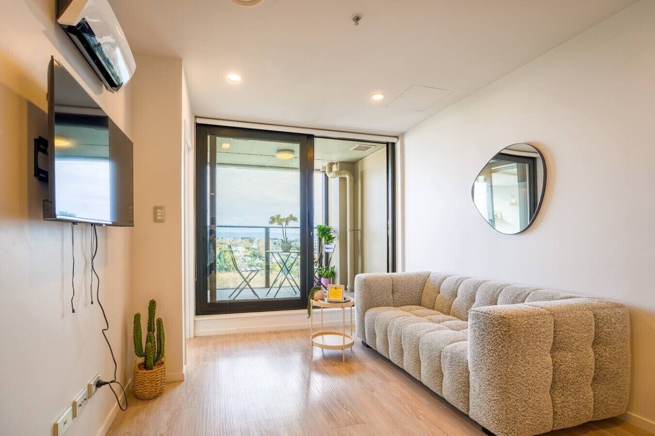 A modern living room with a sofa, a flat-screen TV mounted on the wall, a large window with a view, and a round mirror on the wall.