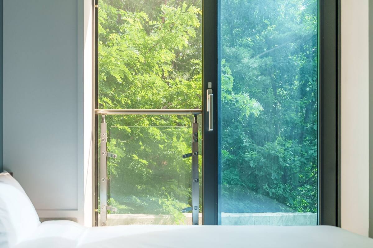 A bedroom with a large window offering a view of dense green foliage.