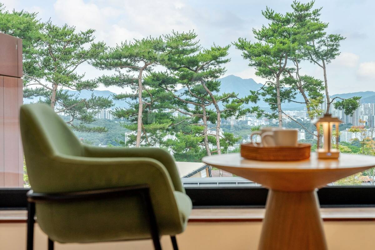 A balcony with two green chairs, a wooden table with a cup and a lantern, and a cityscape view with trees and mountains in the background.