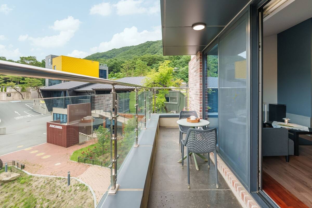 A modern balcony with a glass railing, a small table with chairs, and a view of a green park and buildings.