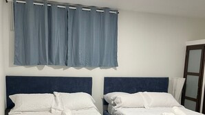 Two beds with dark blue headboards, white linens, and towels, a window with blue curtains, and a white wall. - Apartment 202A (San Andrés Islas)