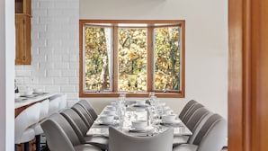 A dining area with a long table set for six, a wooden cabinet, and a window with a view of autumn trees.