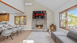 A modern living room with a stone fireplace, a dining area with chairs and a table, and a large window with a view of trees.