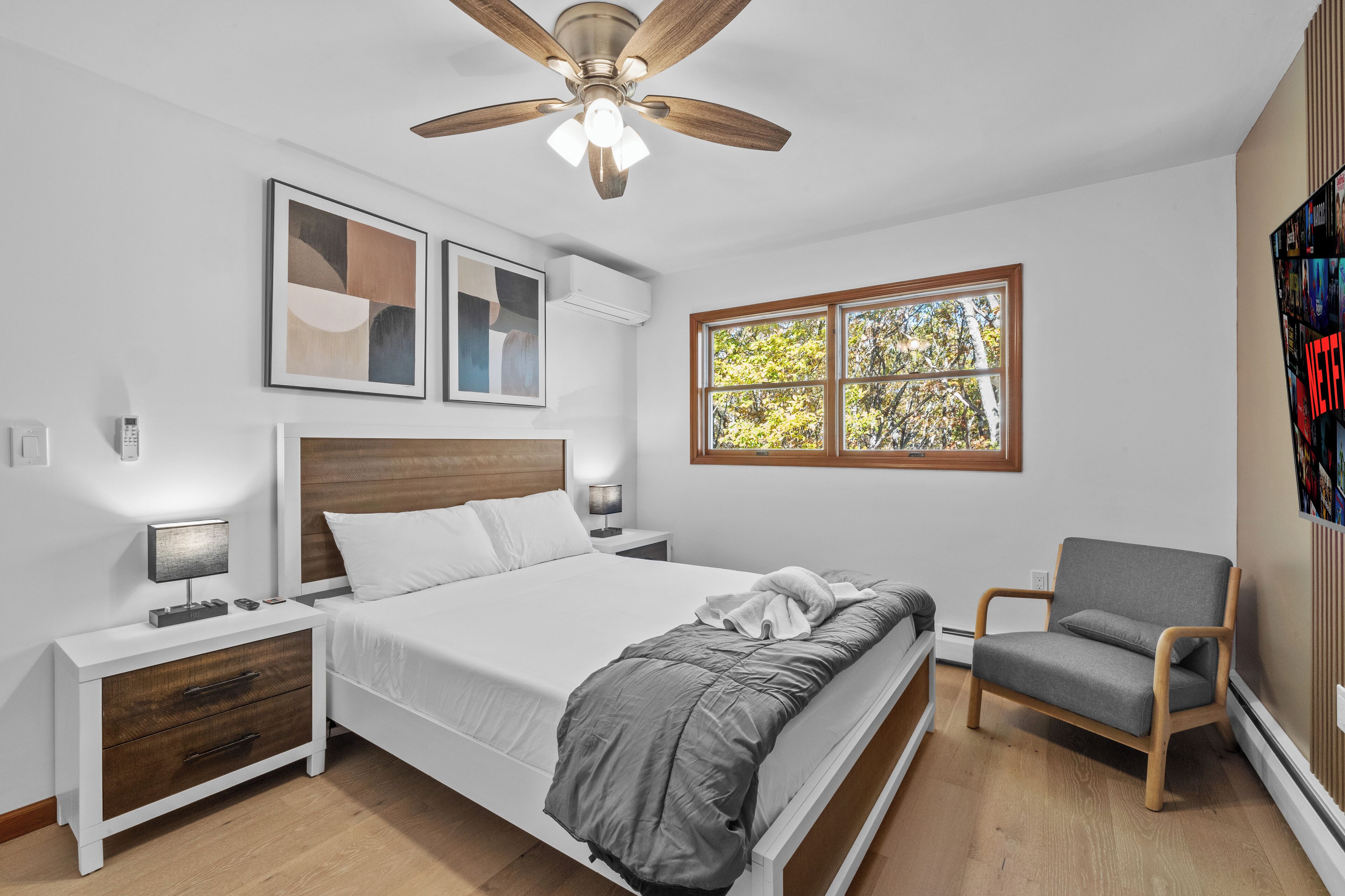 Un dormitorio con cama, mesitas de noche, una silla, ventilador de techo y ventana con vista a árboles.