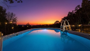 A swimming pool at sunset with a clear sky, surrounded by trees and illuminated by lights.