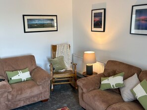 A living room with two brown sofas, a wooden chair, and a table with a lamp. There are framed pictures on the walls.