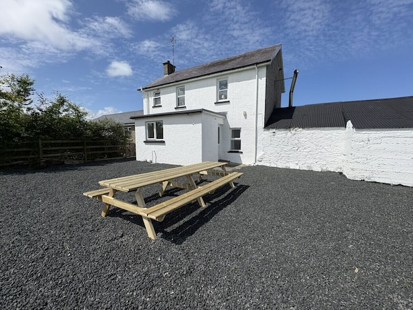 A two-story white house with a gravel driveway and a wooden picnic table in front.