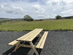 A picnic table in a rural setting with a gravel surface, surrounded by green fields and trees under a cloudy sky.