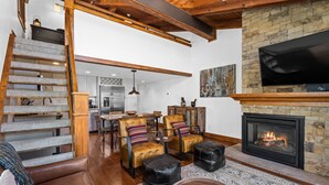 Amplia sala de estar con chimenea, un televisor grande montado en una pared de piedra y una escalera que conduce a un nivel superior.