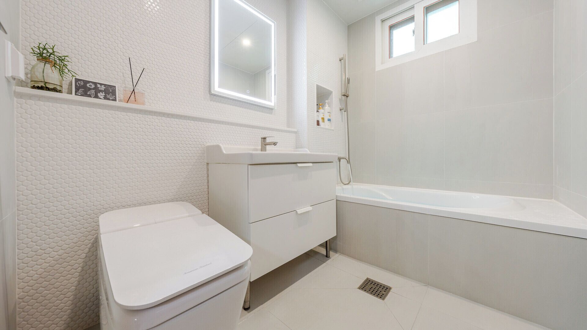 A bathroom with a toilet, sink, and bathtub. There is a shelf with decorative items and a window above the bathtub.