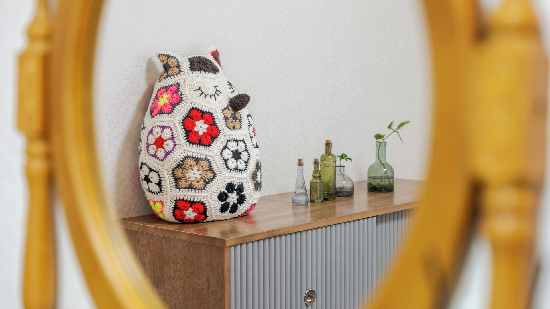 A crocheted bag with a floral pattern on a wooden surface, accompanied by glass bottles and a plant.