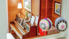 Une chambre d’hôtel avec un piano en bois, une lampe, un jouet en tricot et une valise rouge.
