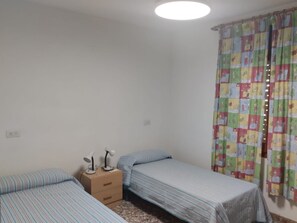 Un petit logement composé d’une seule pièce, comprenant deux lits, une table de chevet avec des lampes et un rideau coloré.