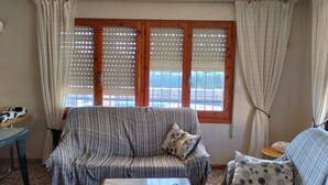 A living room with a sofa, armchair, and coffee table. There are windows with blinds and curtains.