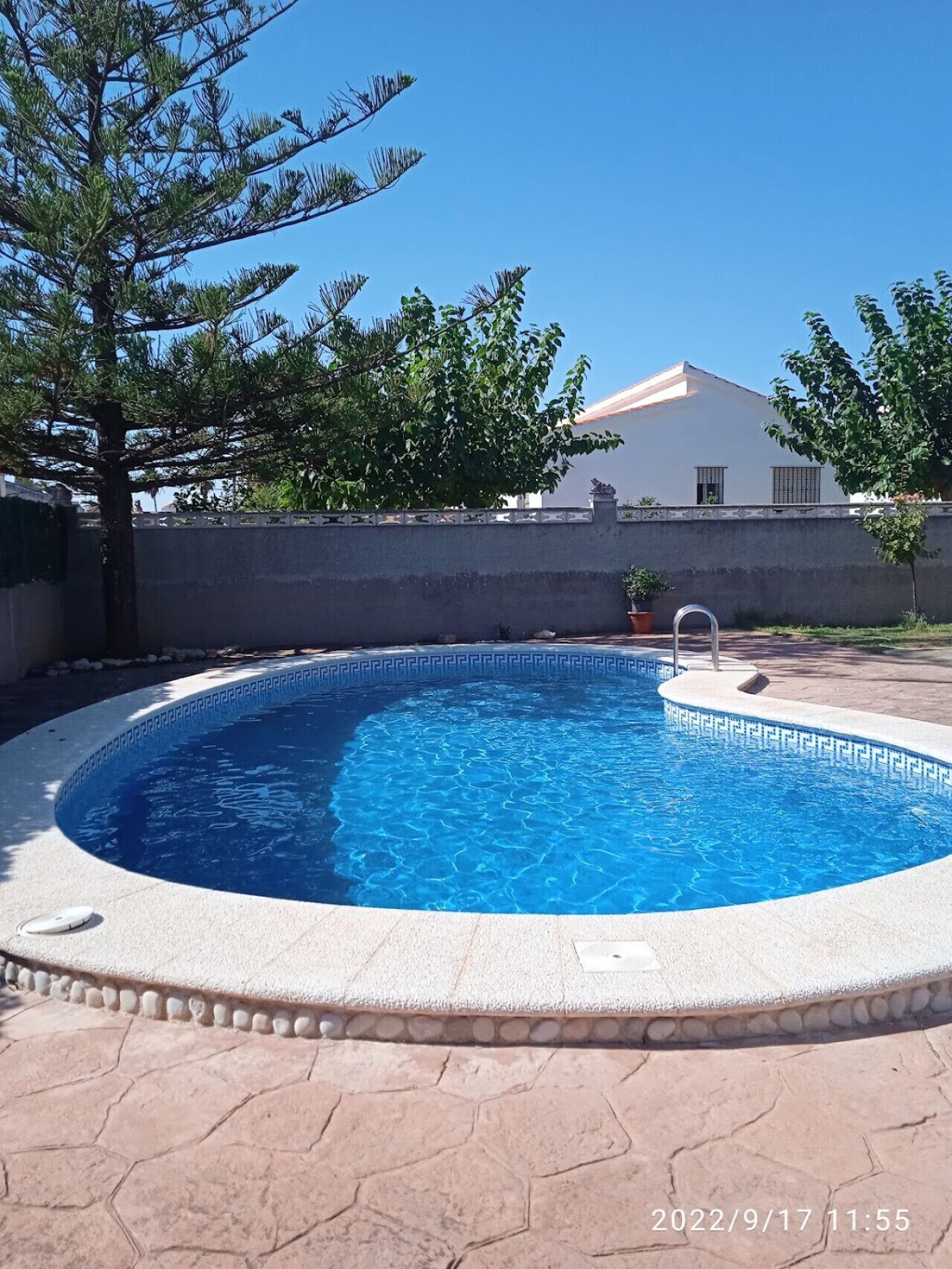A swimming pool with a stone border, surrounded by a paved area and a white wall.
