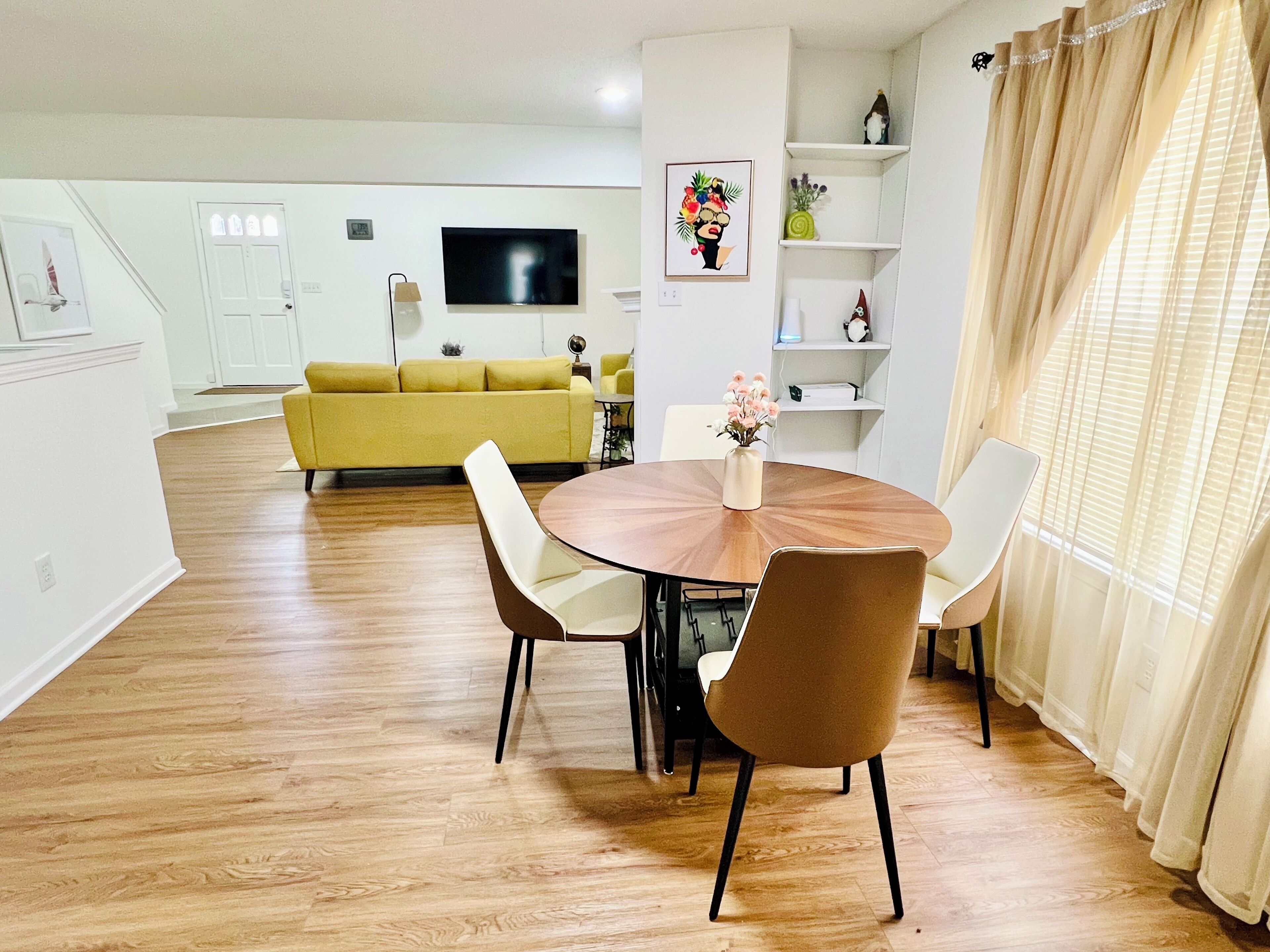 Ein modernes Wohnzimmer mit einem gelben Sofa, einem runden Esstisch mit Stühlen, einem Flachbildfernseher und einer dekorativen Vase.