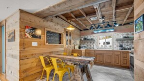 Un interior rústico con paredes de madera, una pizarra con un mensaje de bienvenida, una mesa de comedor de madera con sillas amarillas y una zona de cocina.
