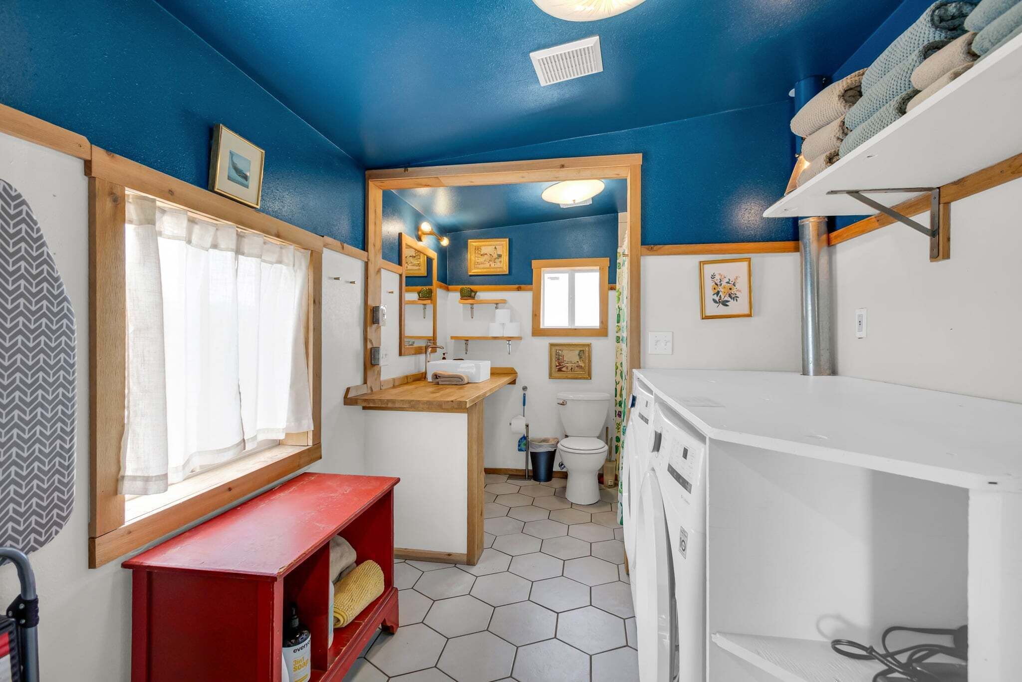 Une petite salle de bain avec un plafond bleu, un meuble-lavabo en bois, un banc rouge, un lavabo blanc et des toilettes.