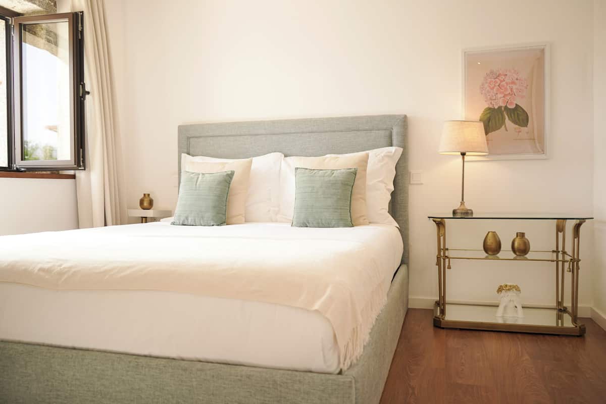 A neatly made bed with a grey headboard, a bedside table with a lamp, and a framed floral artwork on the wall.