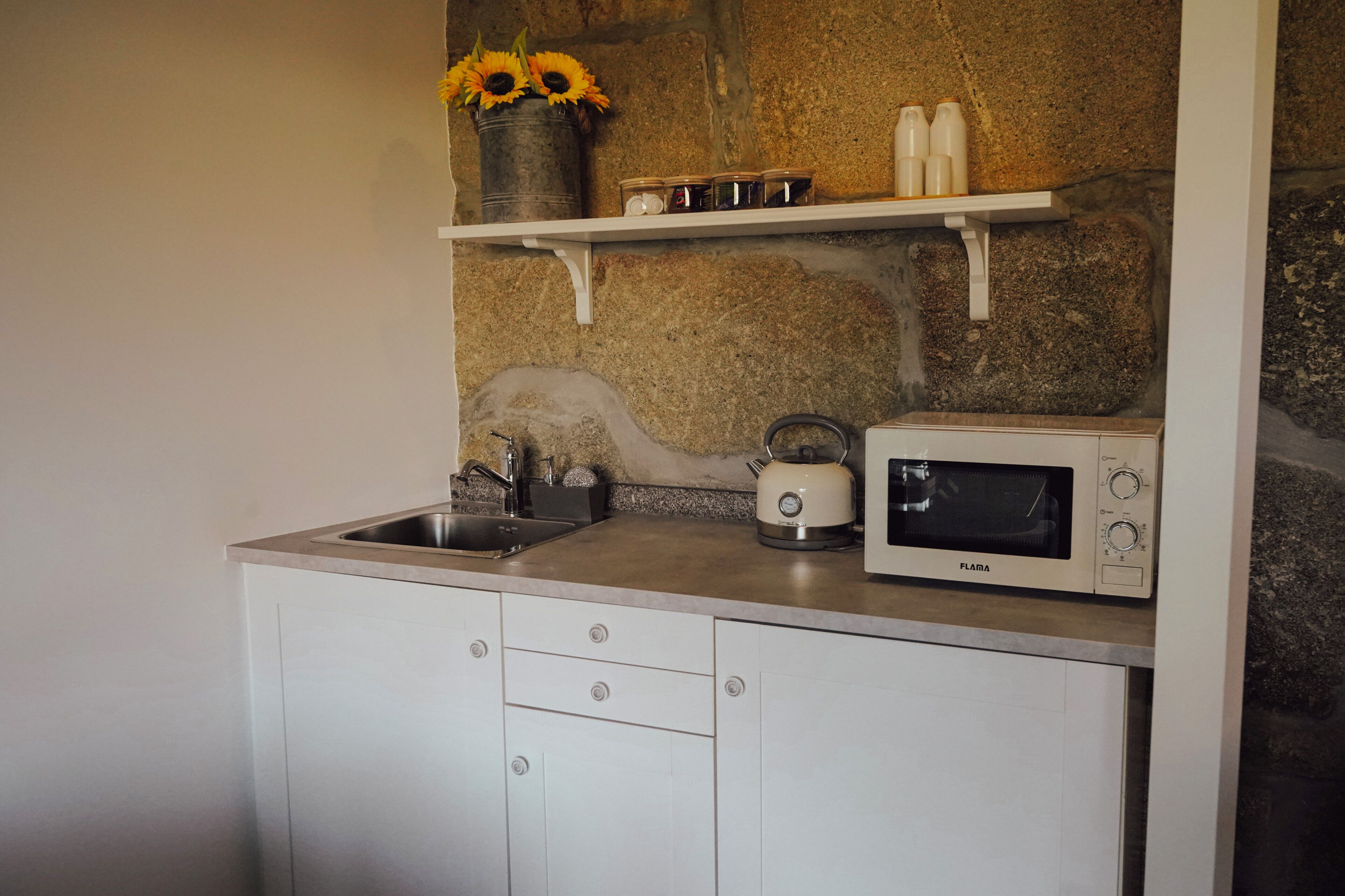 A kitchen with a sink, a toaster, a microwave, and a shelf with jars and a vase of sunflowers.