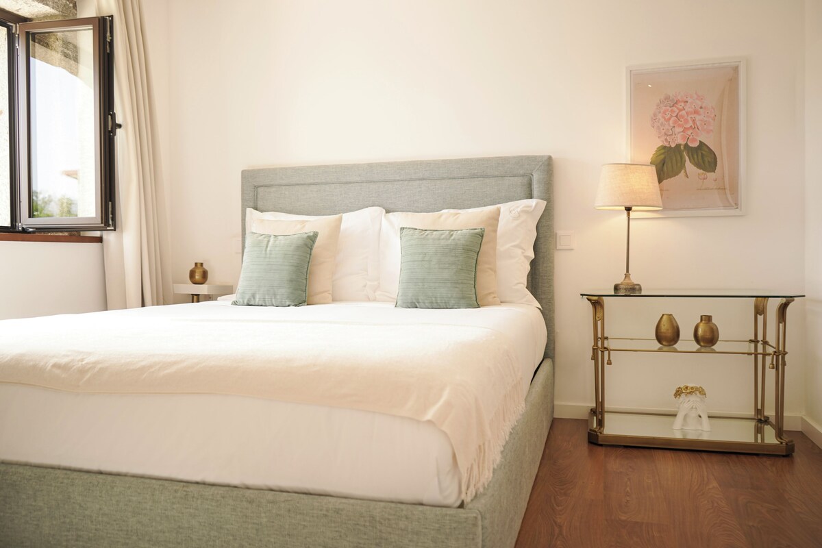 A neatly made bed with a grey headboard, a bedside table with a lamp, and a framed floral artwork on the wall.