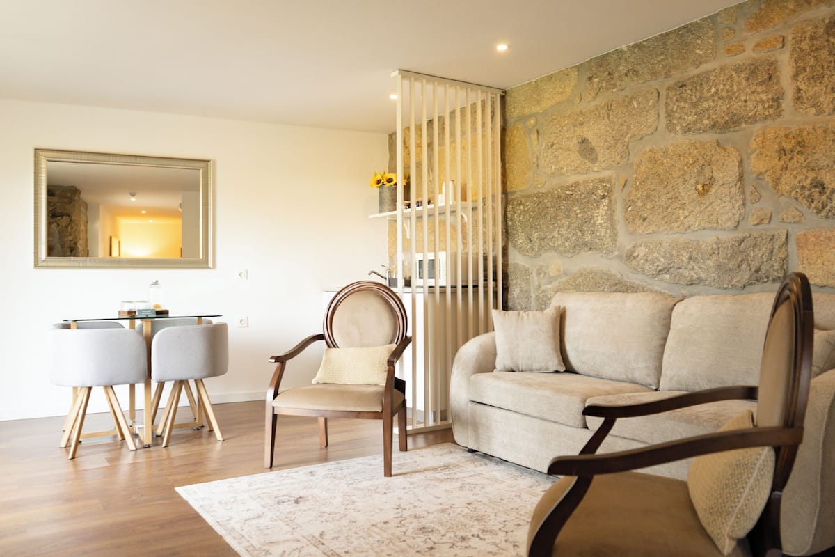 A living room with a stone wall, a sofa, a chair, a glass table with two chairs, and a mirror.
