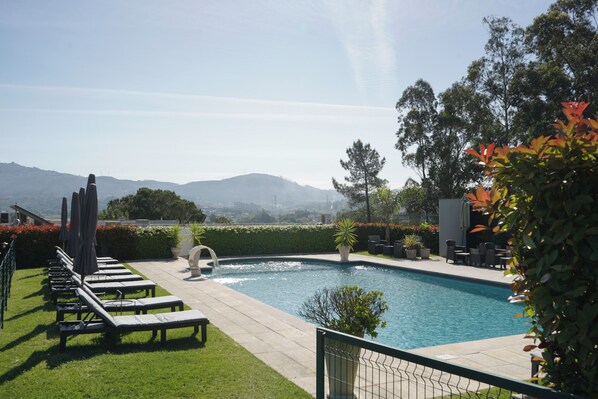 Piscina con sillones de descanso y una fuente, rodeada de vegetación y vistas a colinas distantes.