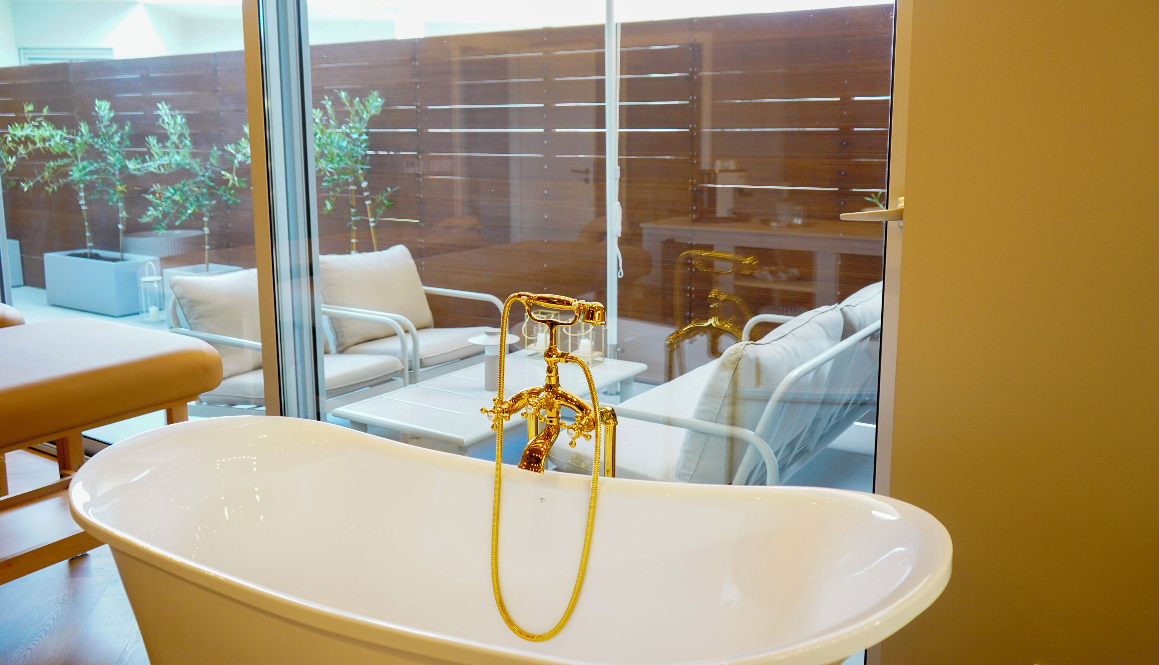 A bathroom with a freestanding bathtub, a glass shower enclosure, and a view of an outdoor seating area with a wooden fence and plants.