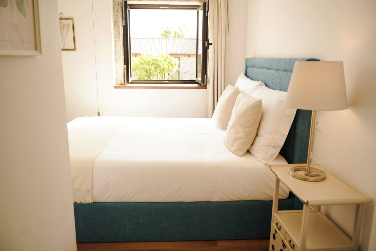 A neatly made bed with a blue headboard, a bedside lamp, and a window with a view of a building and trees outside.