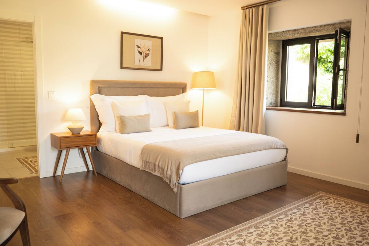 A bedroom with a bed, bedside lamps, a window with curtains, a framed picture on the wall, and a patterned rug.