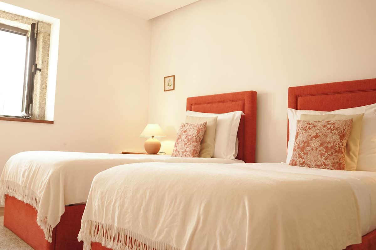 A hotel room with two beds, a window, a lamp, and a small picture on the wall.