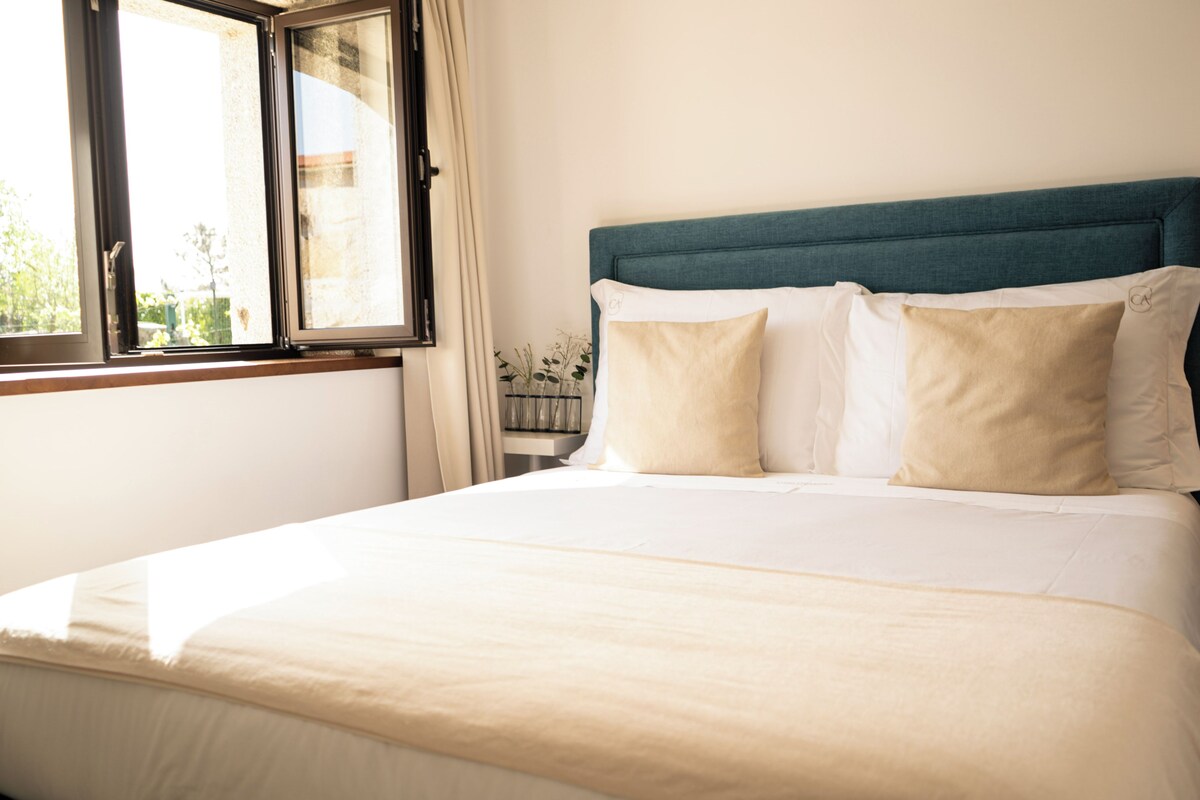 A neatly made bed with a teal headboard, a window with curtains, and a vase with flowers on a nightstand.