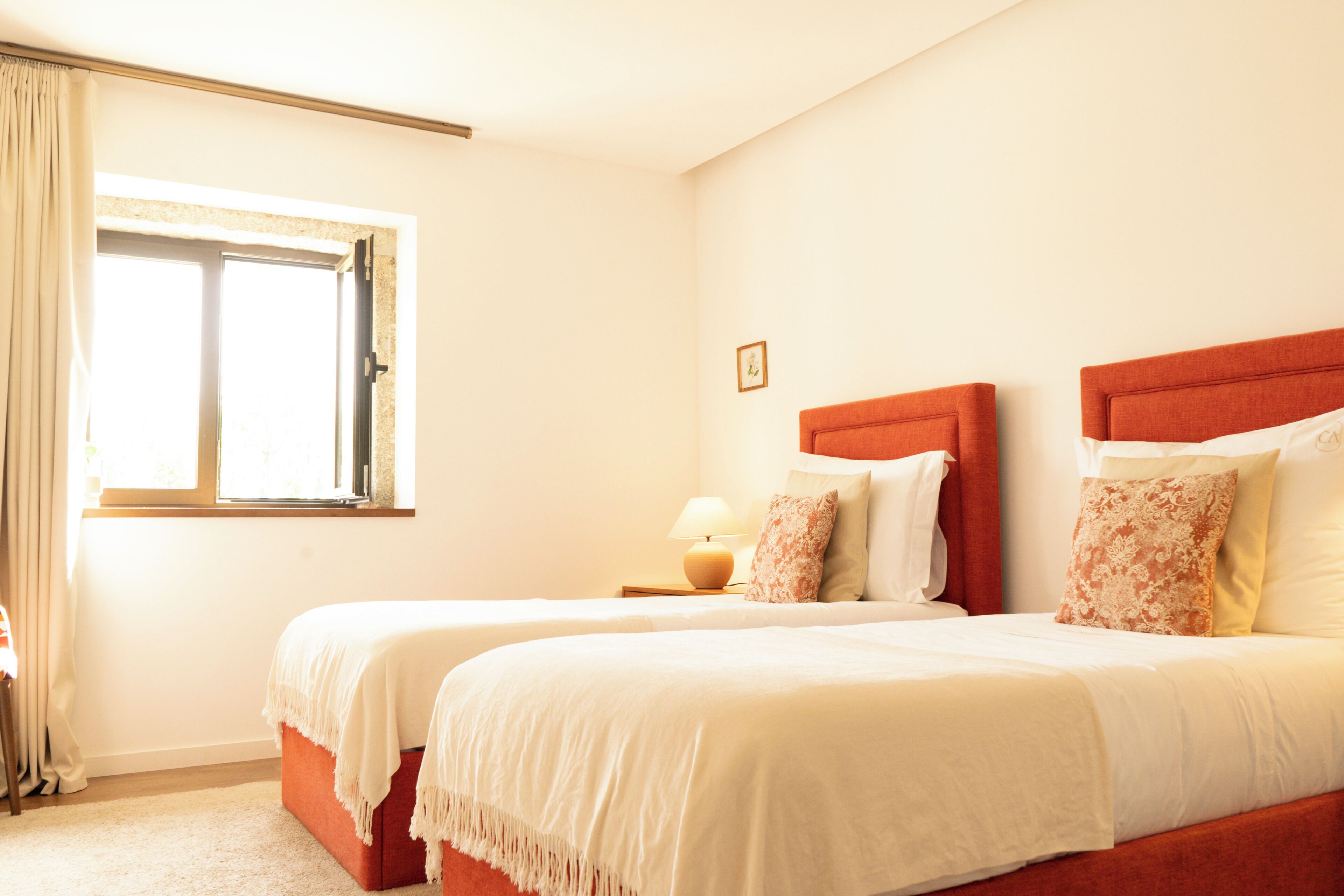 Une chambre d’hôtel avec deux lits, une fenêtre avec des rideaux, une lampe de chevet et un petit tableau encadré accroché au mur.