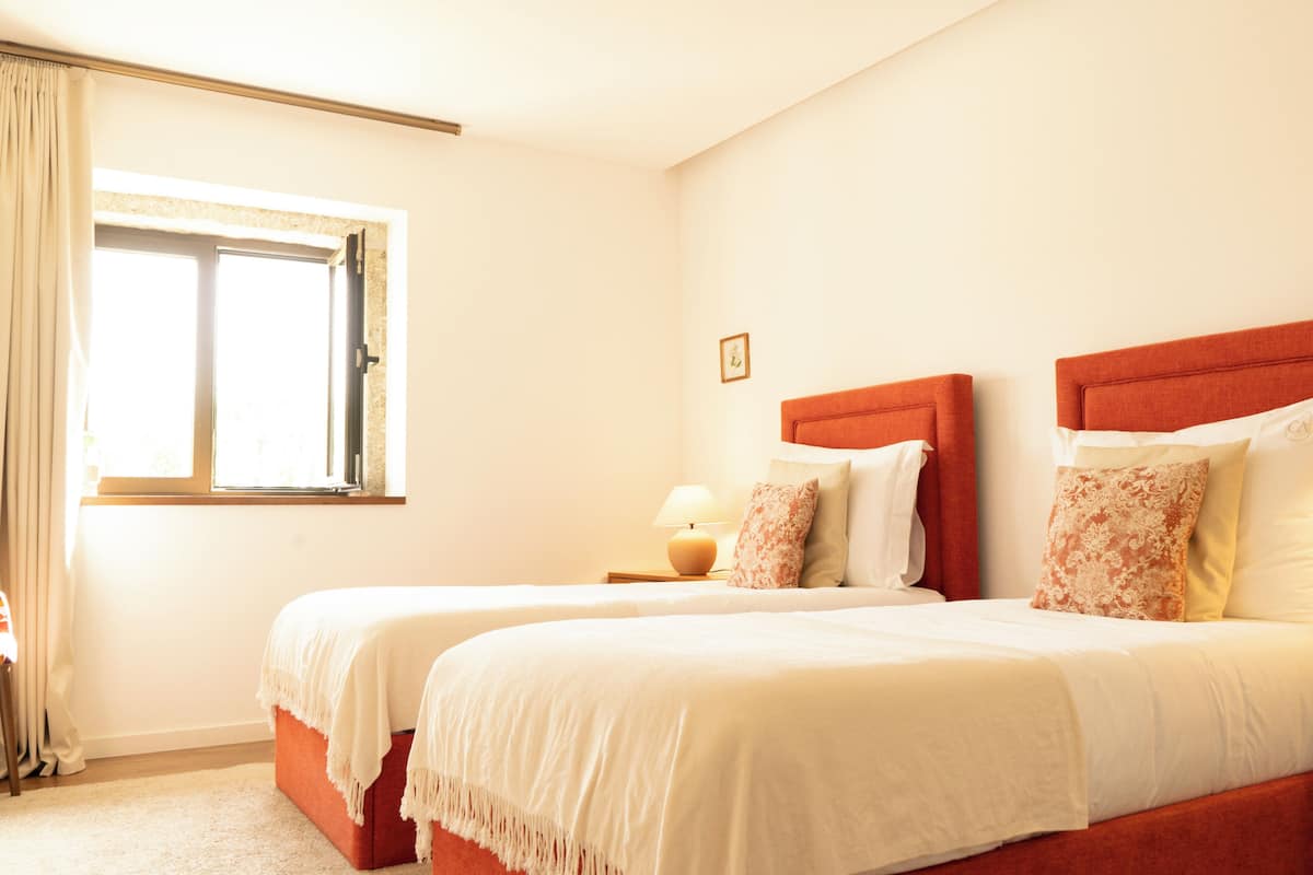 A hotel room with two beds, a window with curtains, a bedside lamp, and a small framed picture on the wall.
